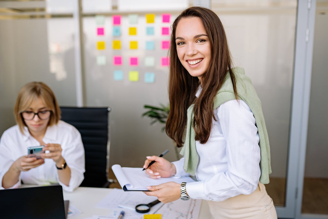 A smiling businesswoman holds a clipboard in a modern office setting.